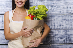 Carrying a healthy bag. Cropped image of beautiful young woman in apron holding paper shopping bag full of fresh vegetables and smiling while standing in front of wooden background